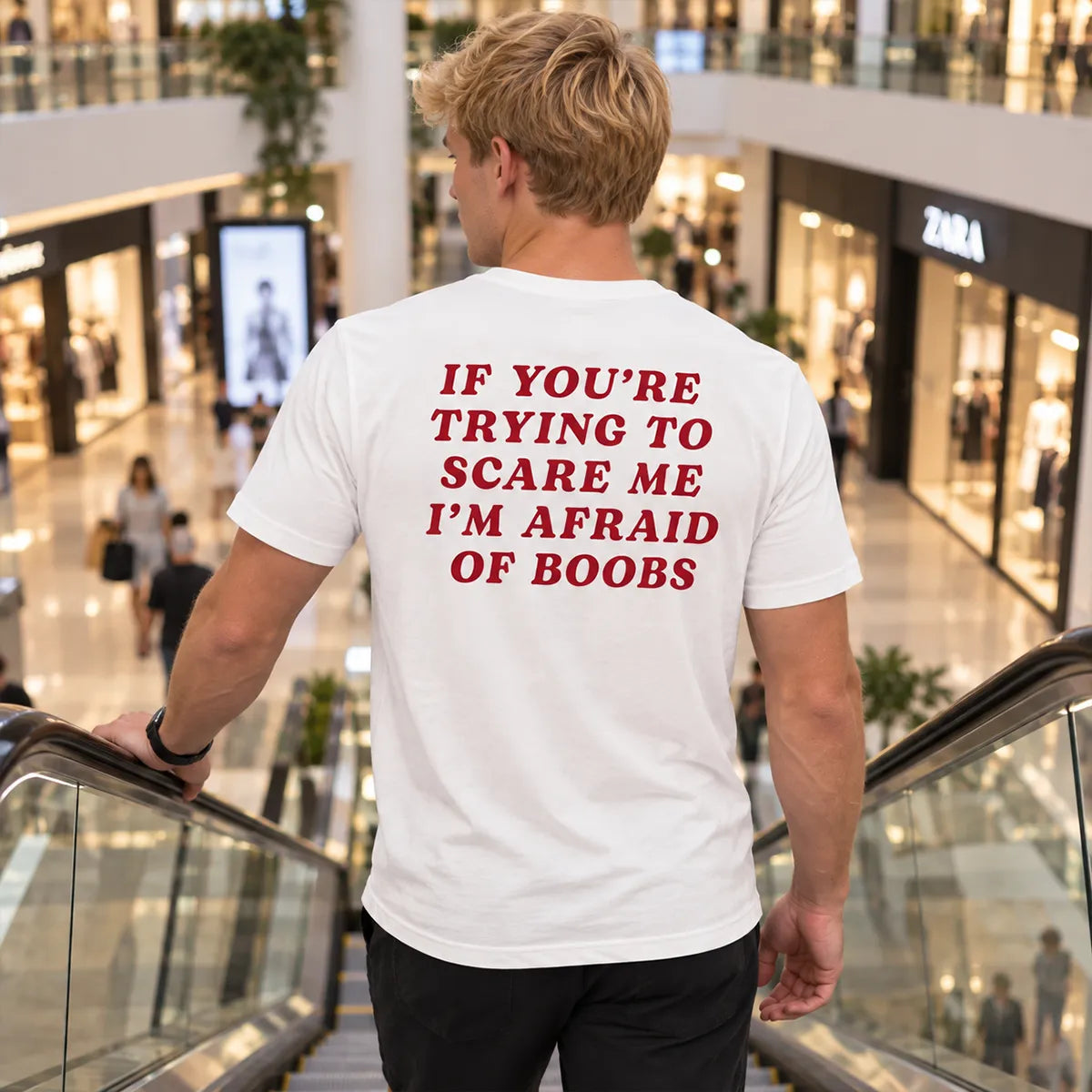 Blonde man riding down an escalator in a mall wearing a white graphic T-shirt with “If You’re Trying to Scare Me I’m Afraid of Boobs” printed on the back.