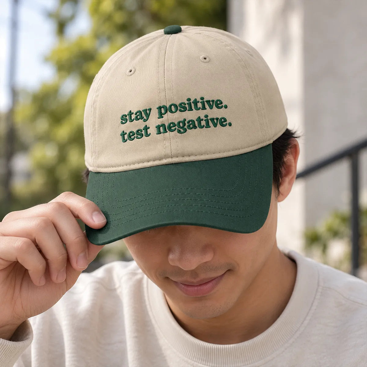 Close-up of an Asian American man wearing a cream and green embroidered cap reading “Stay Positive. Test Negative.”