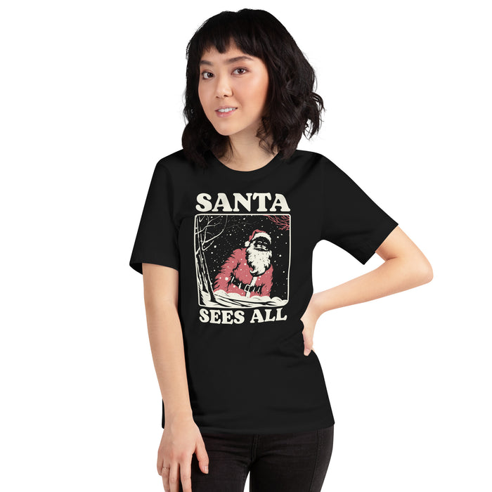 Model wearing the 'Santa Sees All' T-shirt in Black, featuring a vintage-style snowy Santa design and bold text reminding everyone that Santa is watching.