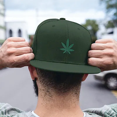 man wearing a snapback hat backwards, with a green cannabis leaf on a spruce colored hat