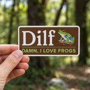 Hand holding a “Dilf, Damn I Love Frogs” sticker with a green frog illustration in front of a softly blurred forest background.