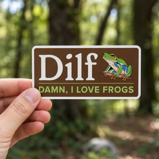 Hand holding a “Dilf, Damn I Love Frogs” sticker with a green frog illustration in front of a softly blurred forest background.