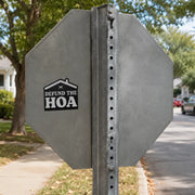 Black “Defund the HOA” house-shaped sticker placed on the gray metal back of a stop sign in a quiet residential neighborhood.