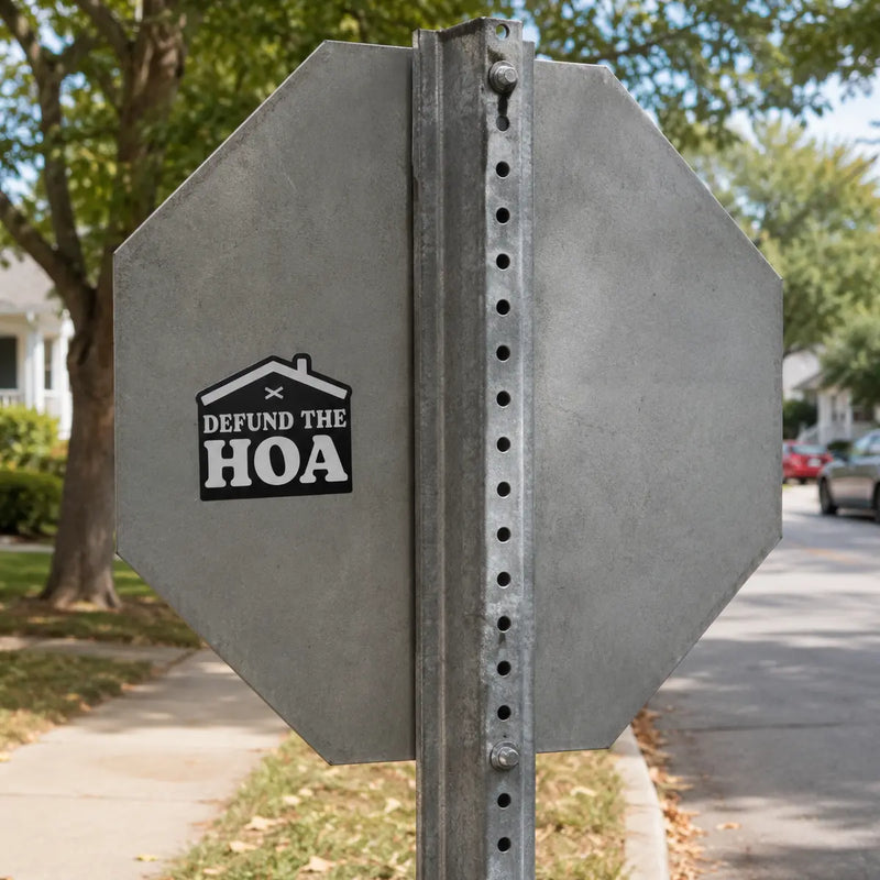 Black “Defund the HOA” house-shaped sticker placed on the gray metal back of a stop sign in a quiet residential neighborhood.