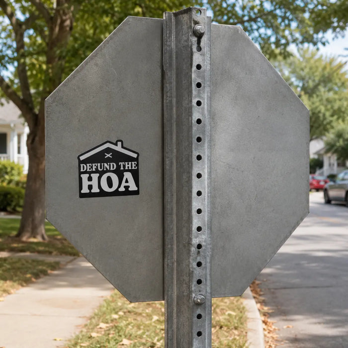 Black “Defund the HOA” house-shaped sticker placed on the gray metal back of a stop sign in a quiet residential neighborhood.