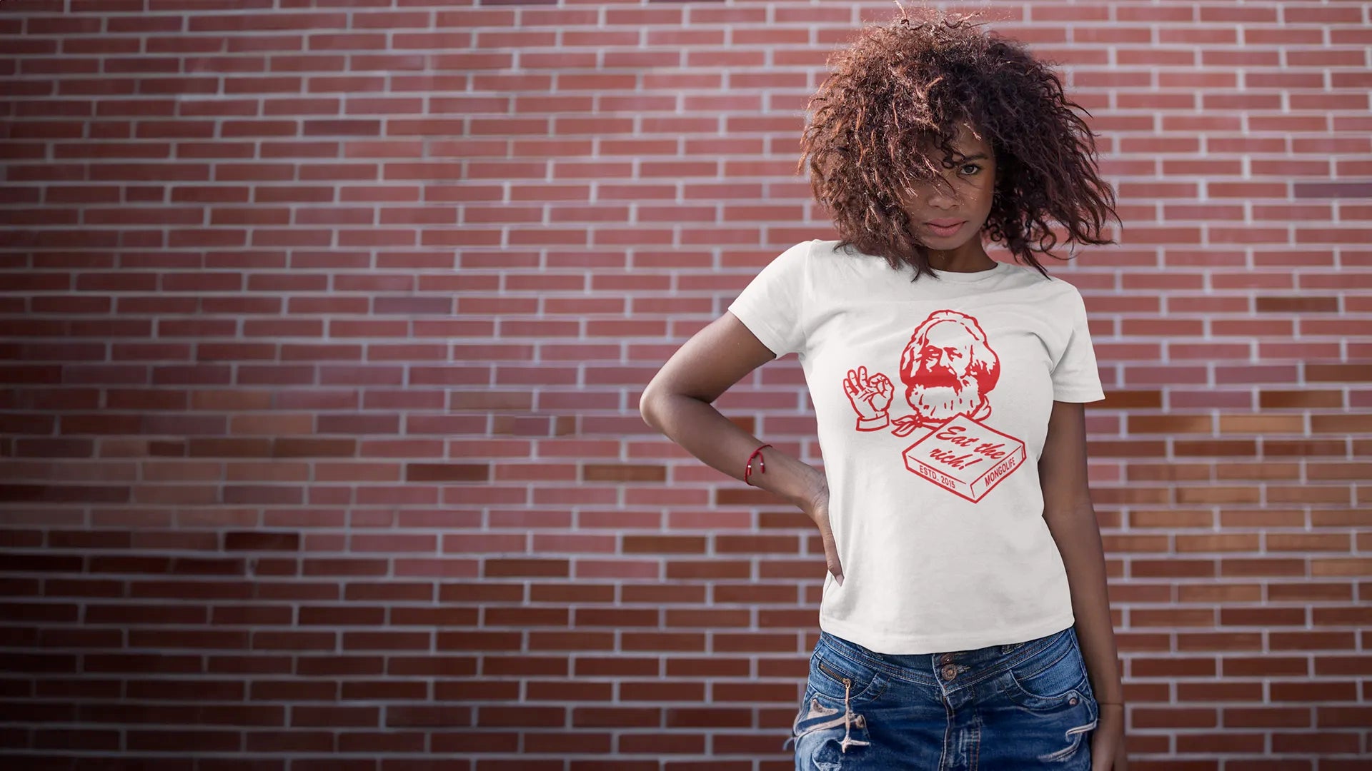 Woman standing in front of a red brick wall, wearing a white t-shirt featuring Karl Marx as a a Pizza Box Chef Drawing