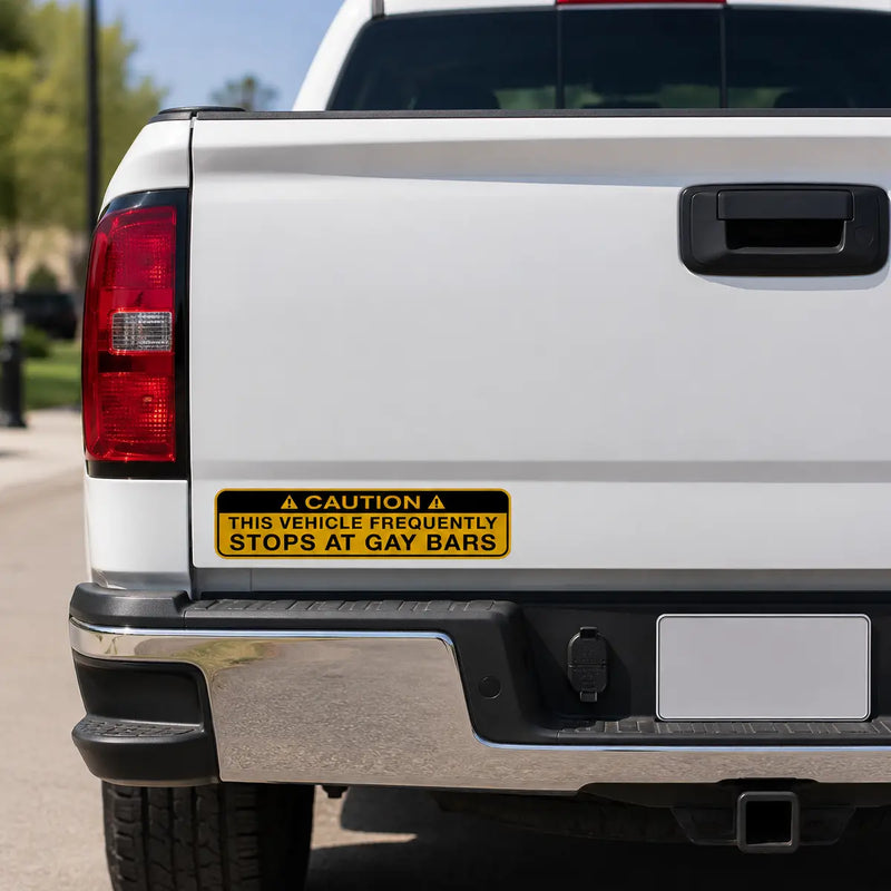 Yellow caution-style prank bumper sticker reading “This vehicle frequently stops at gay bars” on the rear of a white pickup truck.