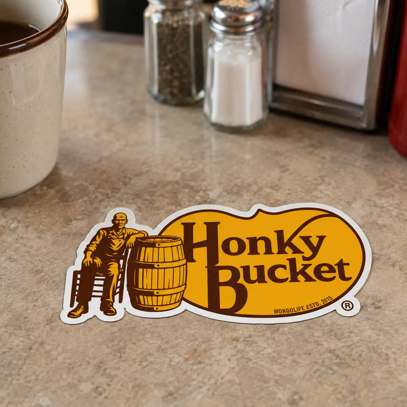Honky Bucket parody sticker placed on a diner table beside a coffee mug, salt and pepper shakers, napkin dispenser, and ketchup bottle.